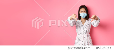 Coronavirus, social distancing and lifestyle concept. Disappointed asian woman in face mask looking judgemental, showing thumbs-down, express negative opinion, pink background 105108053