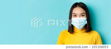 Covid-19, social distancing and pandemic concept. Close up of young asian woman with short dark hair, wearing medical mask and smiling with eyes, looking hopeful at camera 105108055