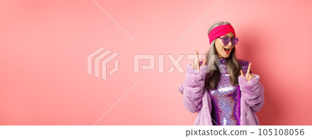 Hipster granny in sunglasses and glittering dress posing for photo with peace, victory signs, standing sassy against pink background 105108056