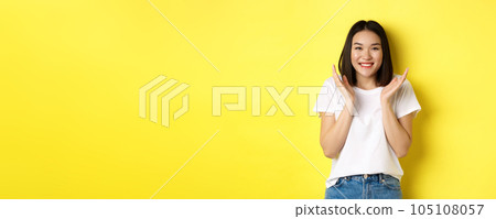 Beauty and fashion concept. Excited asian woman clap hands and smiling happy at camera, standing in white t-shirt against yellow background Beauty and fashion concept. Excited asian woman clap hands and smiling happy at camera, standing in white t-shirt against yellow background 105108057
