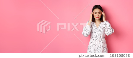 Image of upset asian woman having headache, feeling unwell or dizzy, close eyes and massaging head, suffering migraine, standing over pink background 105108058