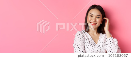 Close-up of beautiful asian woman smiling happy, touching new haircut, standing over pink background 105108067