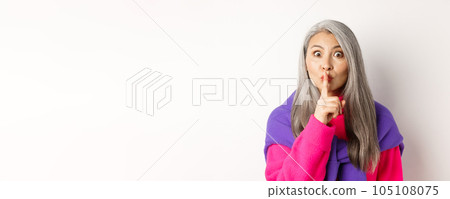 Close-up of stylish senior asian woman in hipster outfit telling hush, shushing at looking at camera, show taboo gesture, standing over white background Close-up of stylish senior asian woman in hipster outfit telling hush, shushing at looking at camera, show taboo gesture, standing over white background 105108075