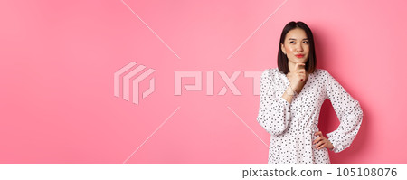 Thoughtful happy asian woman making decision, smiling satisfied and looking at upper left logo, thinking or choosing, standing in dress against pink background Thoughtful happy asian woman making decision, smiling satisfied and looking at upper left logo, thinking or choosing, standing in dress against pink background 105108076