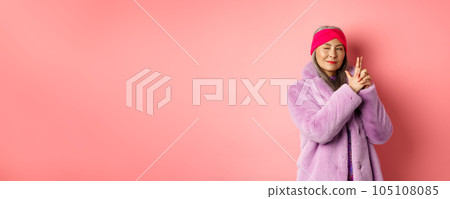 Fashion and shopping concept. Cool and stylish asian senior lady in purple fake fur coat, making finger gun gesture and looking left with sassy smile, acting like secret agent, pink background Fashion and shopping concept. Cool and stylish asian senior lady in purple fake fur coat, making finger gun gesture and looking left with sassy smile, acting like secret agent, pink background 105108085