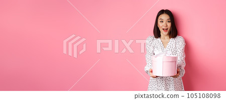 Cute asian woman congratulate with holiday or birthday, giving gift in cute box, standing over pink background 105108098