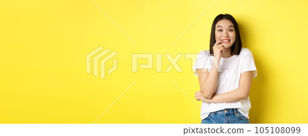 Portrait of cheerful asian girl wants something, looking with desire and temptation at camera, smiling at camera, standing ovr yellow background 105108099