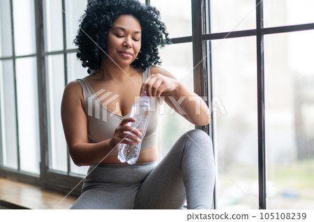 Contented curly-haired woman in sportswear sitting on the wondow with bottle of water 105108239