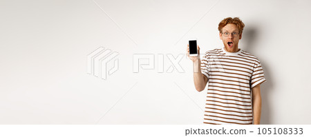 Technology and e-commerce concept. Surprised and shocked redhead guy checking out online promotion, showing blank smartphone screen and drop jaw, white background 105108333