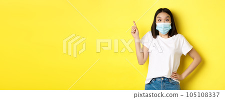 Covid, health care and pandemic concept. Asian female model in medical mask and white t-shirt pointing finger at upper left corner logo, showing promotion, yellow background Covid, health care and pandemic concept. Asian female model in medical mask and white t-shirt pointing finger at upper left corner logo, showing promotion, yellow background 105108337