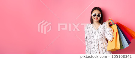 Young asian woman in sunglasses going shopping, holding bags from malls and stores and smiling, standing over pink background 105108341