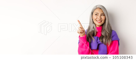 Close-up of beautiful asian senior woman smiling, looking and pointing upper left corner advertisement, checking out promo offer, standing over white background 105108343