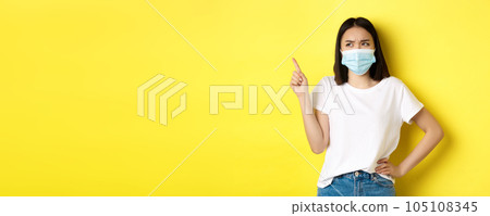 Covid, health care and pandemic concept. Asian woman in medical mask and white t-shirt pointing finger at upper left corner logo, showing promotion, yellow background 105108345