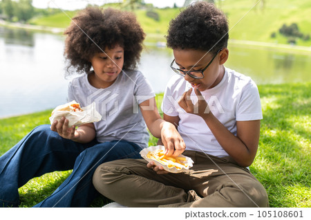 Dark skinned friends or sister and brother sitting on grass in park and eating junk food enjoying. 105108601