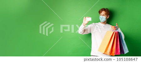 Handsome young man in medical mask, showing plastic credit card and shopping bags with items purchased with discount, green background Handsome young man in medical mask, showing plastic credit card and shopping bags with items purchased with discount, green background 105108647
