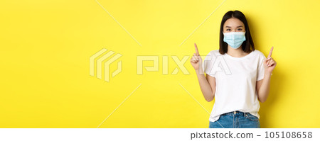 Covid-19, pandemic and social distancing concept. Young asian woman in white t-shirt and medical mask from coronavirus, pointing fingers up, showing special offer Covid-19, pandemic and social distancing concept. Young asian woman in white t-shirt and medical mask from coronavirus, pointing fingers up, showing special offer 105108658