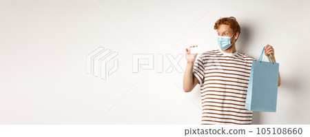 Concept of covid-19 and lifestyle. Cheerful young man with red hair, wear medical mask, showing shopping bag from store and plastic credit card Concept of covid-19 and lifestyle. Cheerful young man with red hair, wear medical mask, showing shopping bag from store and plastic credit card 105108660