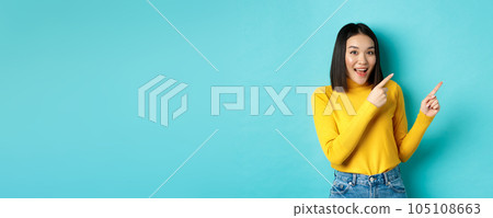Shopping concept. Beautiful chinese girl in yellow sweater pointing fingers at upper right corner logo banner, smiling amused, standing over blue background Shopping concept. Beautiful chinese girl in yellow sweater pointing fingers at upper right corner logo banner, smiling amused, standing over blue background 105108663