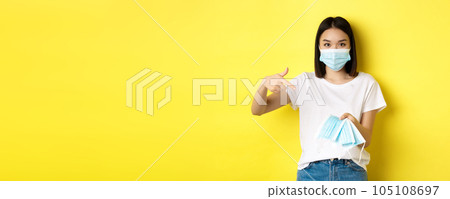 Coronavirus, quarantine and medicine concept. Young asian woman pointing finger at medical masks for going outdoors, standing over yellow background 105108697