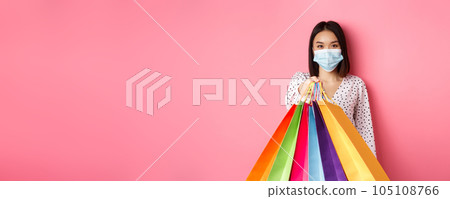 Covid-19, pandemic and lifestyle concept. Beautiful asian woman in face mask giving shopping bags, social distance in store, standing over pink background 105108766
