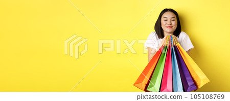 Happy asian female shopper smiling and holding colorful shopping bags, standing against yellow background 105108769