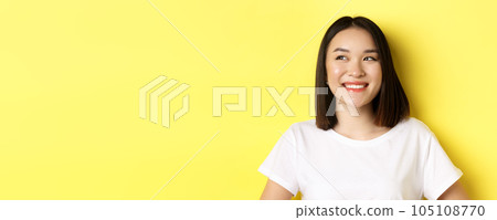 Close up of good-looking asian woman in white t-shirt, smiling and glancing left with pleased face, standing over yellow background 105108770