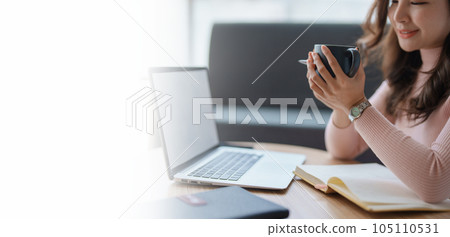 Portrait of a teenage Asian woman using a laptop computer, wearing headphones and using a notebook to study online via video conferencing and holding coffee cup on a wooden desk in living room 105110531