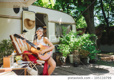 Happy hippie girl is having a good time with playing on guitar in camper trailer. Holiday, vacation, trip concept. Happy hippie girl is having a good time with playing on guitar in camper trailer. Holiday, vacation, trip concept. 105110799