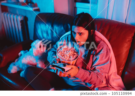 A gamer or a streamer girl at home in a dark room with a game controller and popcorn bucket playing with her dog and sits in front of a monitor or TV. 105110994