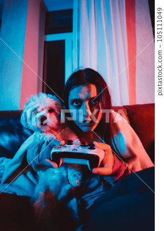 Close up portrait of A gamer or a streamer girl at home in a dark room with a game controller playing with her dog and sits in front of a monitor or TV. 105111049