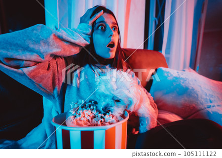 Close up portrait of Beautiful young girl at home in a dark room with a popcorn bucket playing with her dog and sits in front of a monitor or TV. 105111122