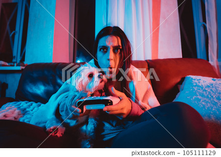 Close up portrait of A gamer or a streamer girl at home in a dark room with a game controller playing with her dog and sits in front of a monitor or TV. 105111129