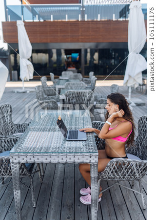 Cute young woman in swimsuit working on laptop sitting on terrace on the beach 105111176