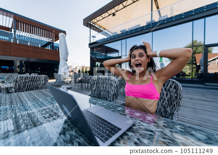 Cute young woman in swimsuit is shocked working on laptop sitting on terrace on the beach 105111249