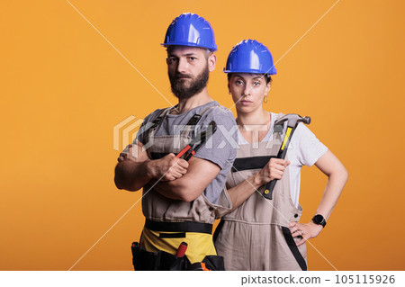 Serious construction workers posing with pair of pliers and hammer, holding slegdehammer and renovating tools in studio. Man and woman working as constructors with overalls and helmet. Serious construction workers posing with pair of pliers and hammer, holding slegdehammer and renovating tools in studio. Man and woman working as constructors with overalls and helmet. 105115926
