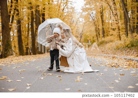 Young woman standing in autumn forest. Blonde woman kissing her daughter. Girl wearing beige sweater and white hat and holding a transparent umbrella. 105117402