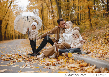 Young man, woman and their children sitting outside in autumn forest. Blond mother and brunette father hugging. Little girl holding a transparent umbrella. 105117403