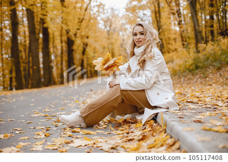 Young woman sitting on a curb in autumn forest. Blonde woman holding a yellow leaves. Girl wearing beige coat and brown trousers. Young woman sitting on a curb in autumn forest. Blonde woman holding a yellow leaves. Girl wearing beige coat and brown trousers. 105117408