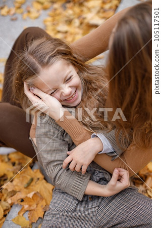 Young woman and little girl in autumn forest. Woman hugging her daughter. Girl wearing fashion clothes and lying on mother's lap. Young woman and little girl in autumn forest. Woman hugging her daughter. Girl wearing fashion clothes and lying on mother's lap. 105117501