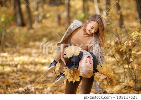 Young woman and little boy in autumn forest. Woman holding her son on her hands. Boy wearing fashion clothes and holding yellow leaves. 105117523