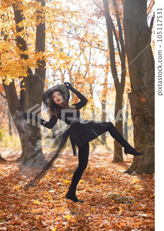 Brunette woman in witch costume standing in autumn forest on Halloween day. Woman wearing black clothes and cone hat. Woman posing for a photo. 105117531