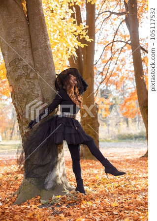 Brunette woman in witch costume standing in autumn forest on Halloween day. Woman wearing black clothes and cone hat. Woman posing for a photo. 105117532