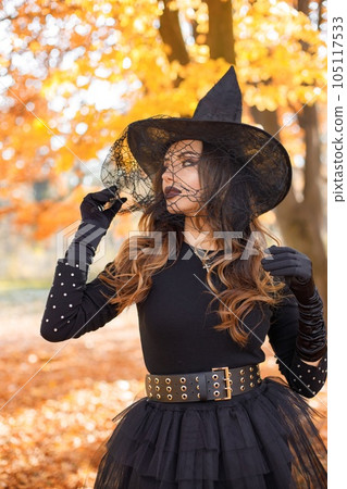 Brunette woman in witch costume standing in autumn forest on Halloween day. Woman wearing black clothes and cone hat. Woman posing for a photo. Brunette woman in witch costume standing in autumn forest on Halloween day. Woman wearing black clothes and cone hat. Woman posing for a photo. 105117533