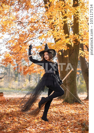 Brunette woman in witch costume standing in autumn forest on Halloween day. Woman wearing black clothes and cone hat. Woman sitting on a broom. 105117534