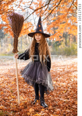 Cute happy little girl dressed in witch costume standing with over autumn forest background. Girl wearing dress and cone hat and holding a broom. Girl dressed for Halloween. 105117553