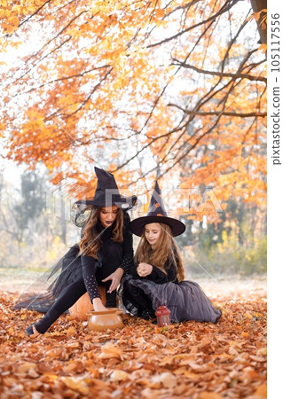 Woman with little girl sitting on a pumpkin in autumn forest. Mother play with her daughter. Mother and daughter wearing witches Halloween costumes and cone hats. 105117556