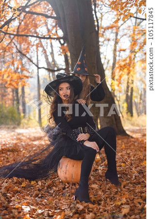 Woman sitting on a pumpkin and little girl standing in autumn forest. Mother play with her daughter. Mother and daughter wearing witches Halloween costumes and cone hats. 105117845