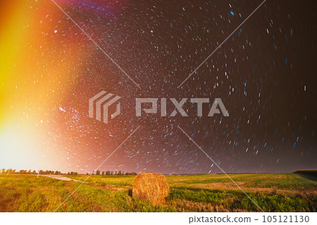 Night Starry Sky in rotation Above Country Field Meadow With Hay Bale After Harvest. Glowing Stars Above Rural Landscape. Colorful flare on sky. Agricultural Landscape under Starry Sky. Colored bright Night Starry Sky in rotation Above Country Field Meadow With Hay Bale After Harvest. Glowing Stars Above Rural Landscape. Colorful flare on sky. Agricultural Landscape under Starry Sky. Colored bright 105121130
