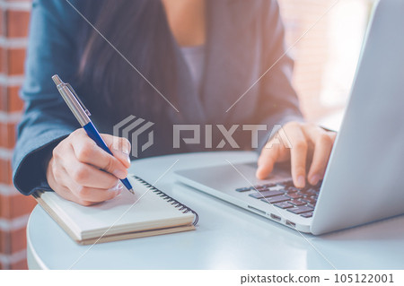 Woman hand is writing on a notepad with a pen and have a laptop computer on the desk in the office. Woman hand is writing on a notepad with a pen and have a laptop computer on the desk in the office. 105122001