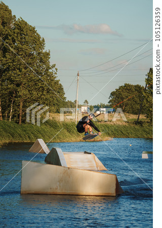 Beautiful view of young man rider holding rope and making extreme jump on wakeboard. Wakeboarding and water sports activity. Low angle shot of man wakeboarding on a lake. Man water skiing at sunset. 105126359